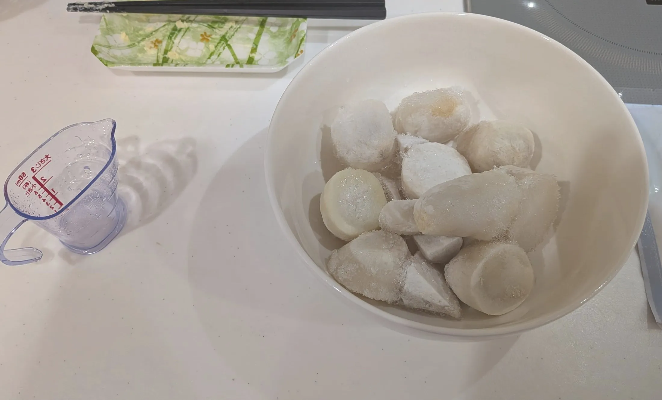 白いボウルに入った凍ったままの里芋。左に水を入れた計量カップ